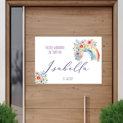 Personalisierbares Banner mit Regenbogen- und Blumenmotiv an einer Holztür befestigt. Text: „Herzlich willkommen zur Taufe von Isabella – 25. Juli 2027“. Außenansicht zur Veranschaulichung des Einsatzes im Außenbereich.