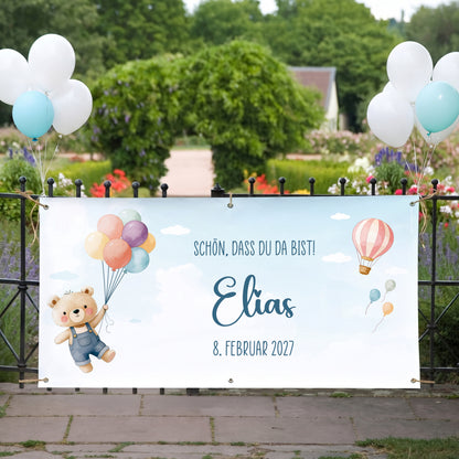 Willkommensbanner zur Geburt eines Jungen mit Name Elias, Teddy Motiv und Datum, befestigt am Gartenzaun mit blauen und weißen Ballons