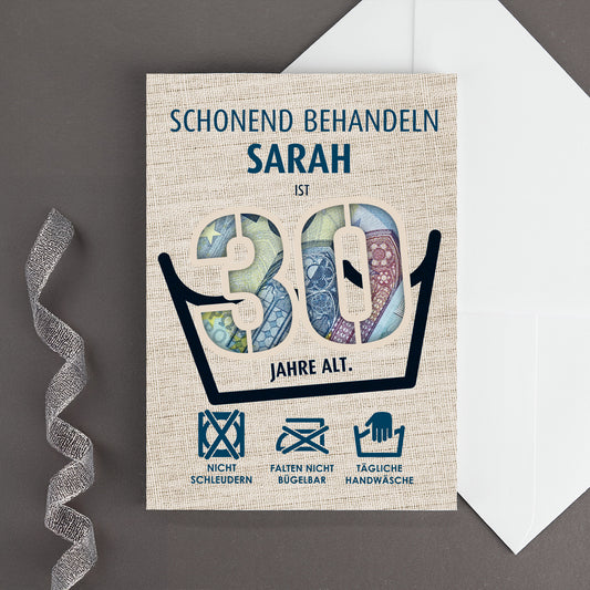 Klappkarte zum 30. Geburtstag mit dem Namen Sarah und ausgestanzter 30, hinterlegt mit Euro-Scheinen, Wäschezeichen und weißem Umschlag auf grauem Hintergrund
