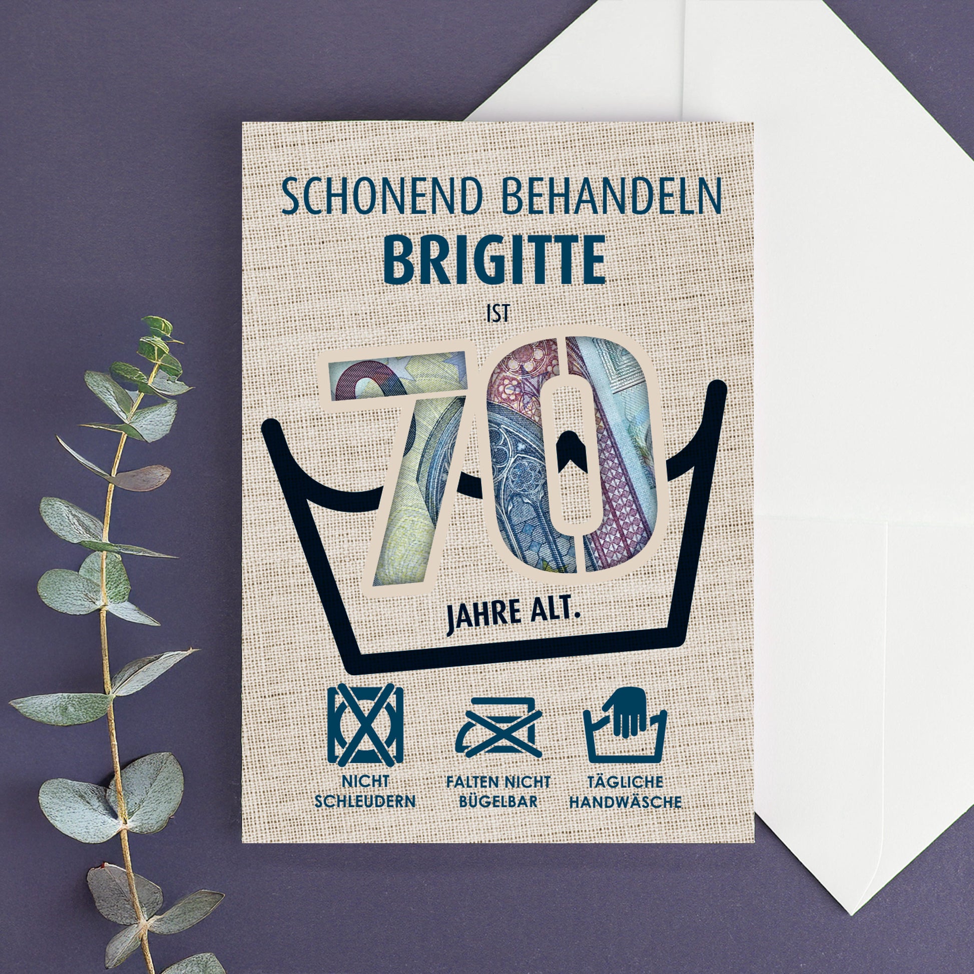 Klappkarte zum 70. Geburtstag mit Geldschein-Hintergrund und dem Namen Brigitte, dekorativ auf lila Untergrund mit Eukalyptuszweig arrangiert