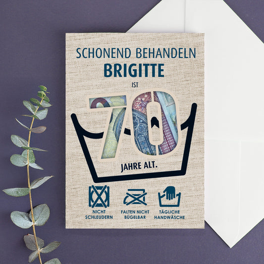 Klappkarte zum 70. Geburtstag mit Geldschein-Hintergrund und dem Namen Brigitte, dekorativ auf lila Untergrund mit Eukalyptuszweig arrangiert