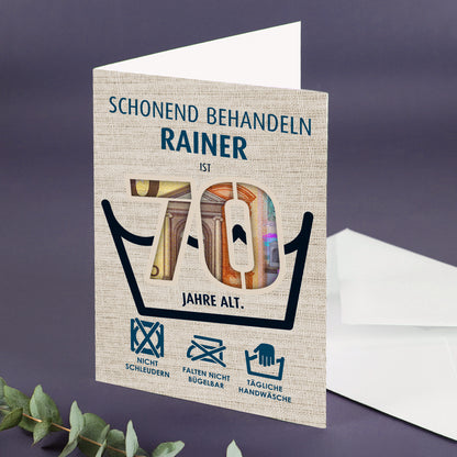Geldgeschenk-Klappkarte mit Zahl 70 und dem Namen Rainer, aufgeklappt auf lila Untergrund neben weißem Umschlag und Eukalyptuszweig