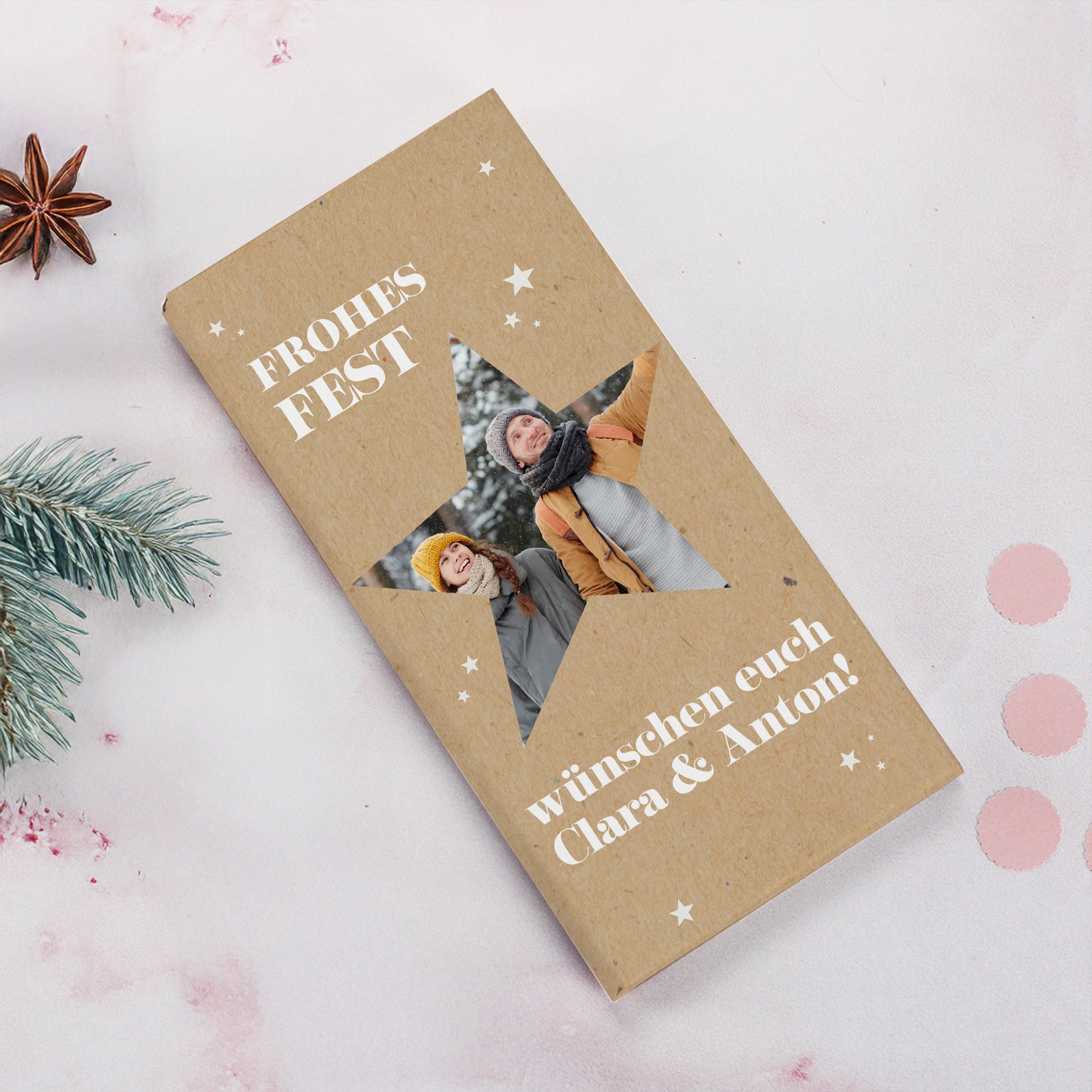 Personalisierte Schokolade zu Weihnachten mit Foto im Sternmotiv. Auf der naturfarbenen Verpackung steht „Frohes Fest“ in weißer Schrift. Im Sternfenster ist ein Foto eines Paares im Schnee zu sehen. Darunter personalisierter Wunschtext. Dekoriert mit Tannenzweig, Sternanis und rosafarbenem Hintergrund.