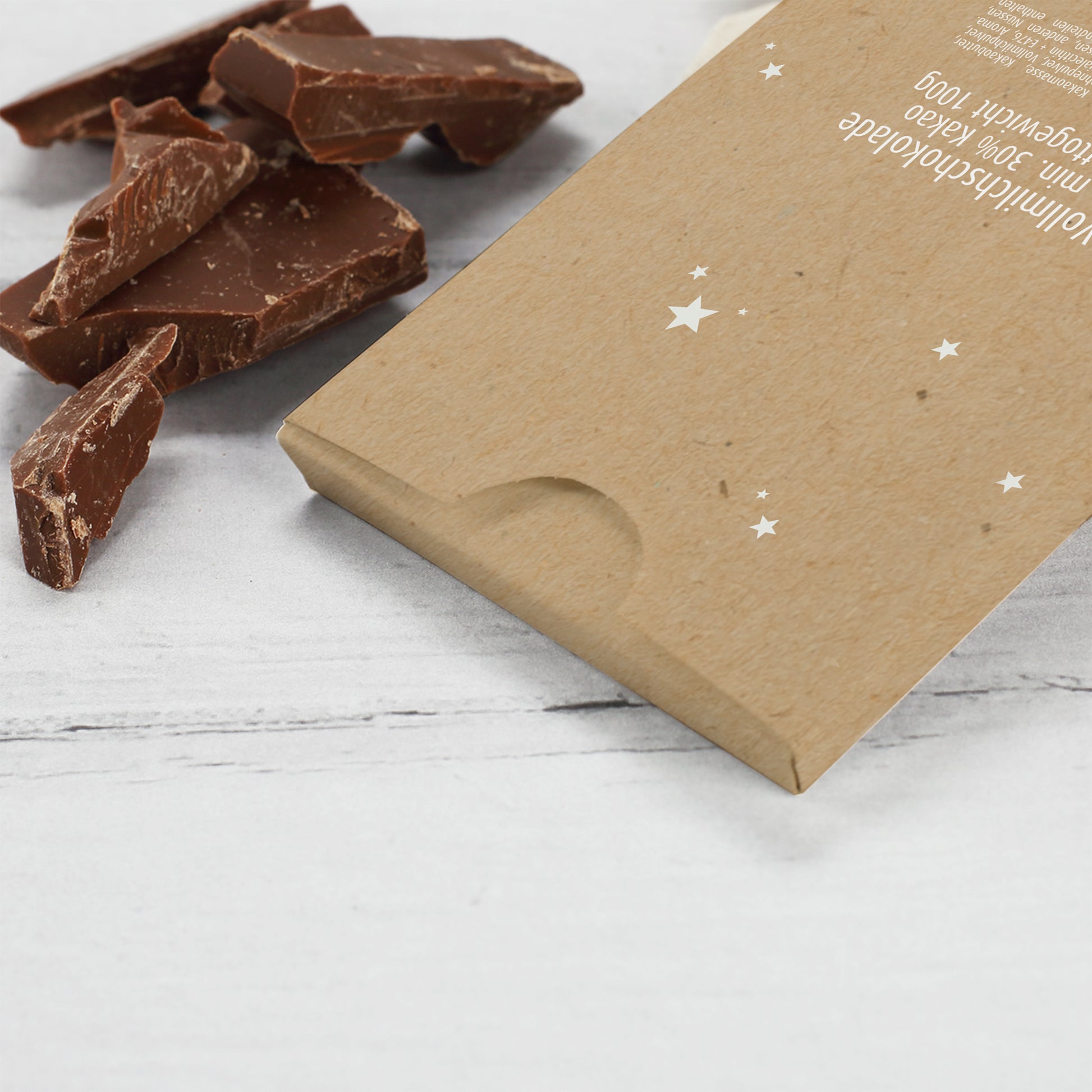 Rückseite der personalisierten Weihnachts-Schokolade mit Sternen-Design auf braunem Karton. Neben der Verpackung liegen Stücke zarter Vollmilchschokolade auf hellem Untergrund. Detailaufnahme des hochwertigen Drucks und der sauberen Verarbeitung.