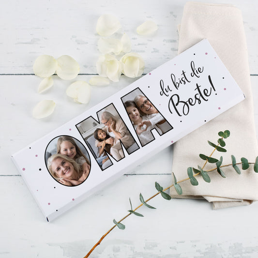 Geschenk-Schokoladentafel XXL mit Fotos im Schriftzug „OMA“ und Rosenblättern als Dekoration auf weißem Tisch