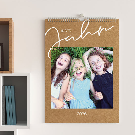 Foto-Wandkalender 2026 in Kraftpapier-Optik, aufgehängt an einer hellen Wand, mit Foto von drei lachenden Kindern und dem Titel „Unser Jahr“.
