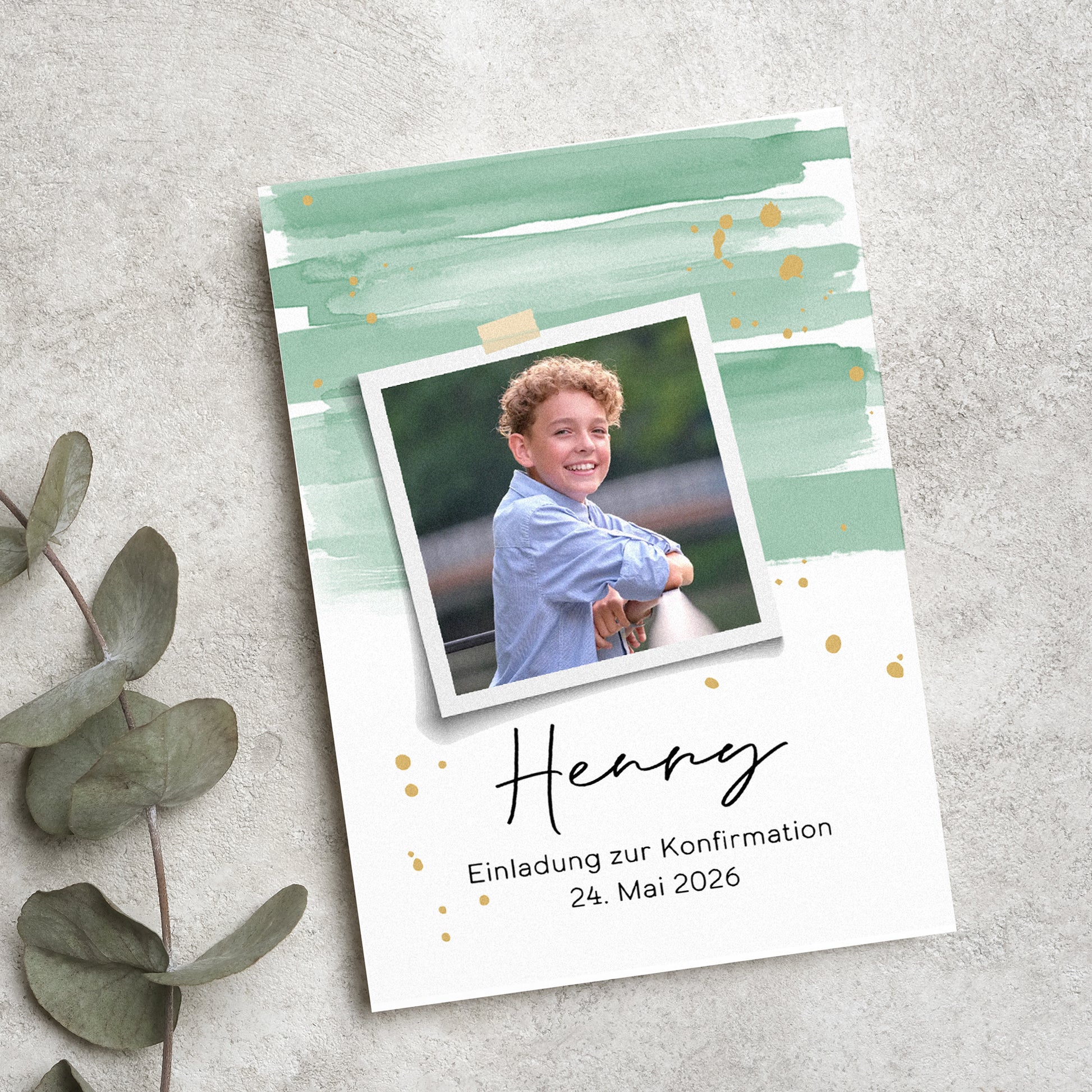 Personalisierte Einladungskarte zur Konfirmation mit modernem Aquarell-Design in Grün und Gold. Ein Polaroid-Foto des Konfirmanden ist mittig platziert, darunter sein Name und das Datum der Feier. Die Karte liegt auf einer strukturierten Steinoberfläche, umgeben von Eukalyptusblättern.