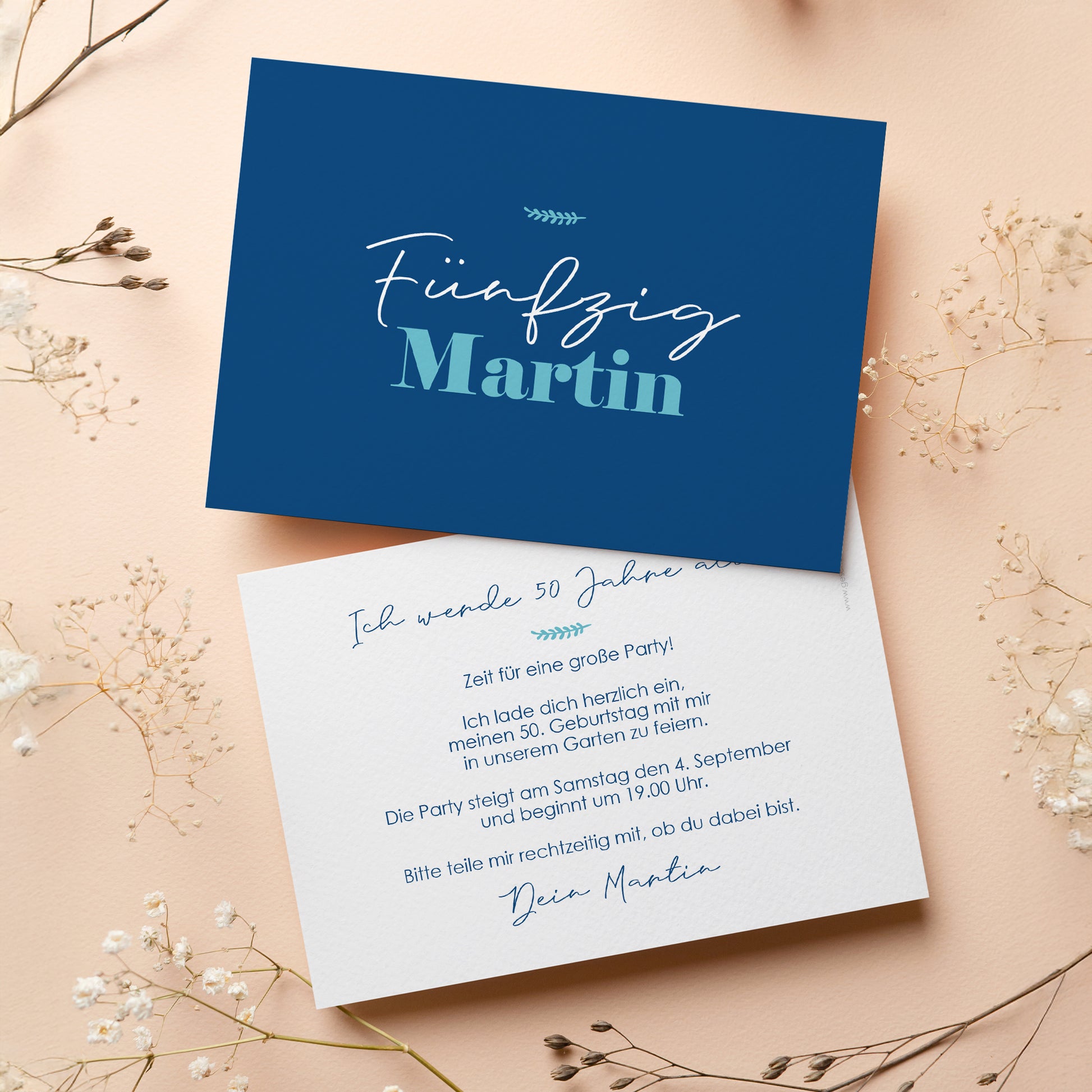 Personalisierte Einladungskarte zum 50. Geburtstag mit stilvollem Design in Dunkelblau. Die Vorderseite zeigt den Schriftzug „Fünfzig Martin“ in einer eleganten Schriftart. Die Rückseite bietet Platz für individuellen Einladungstext und Details zur Feier. Die Karte liegt auf einer beigen Oberfläche mit dekorativen Trockenblumen, passend zur festlichen Atmosphäre.