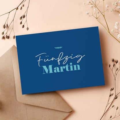 Einladungskarte zum 50. Geburtstag in stilvollem Dunkelblau. Die Vorderseite zeigt den Schriftzug „Fünfzig Martin“ in einer eleganten, modernen Typografie. Die Karte liegt auf einem beigen Untergrund mit einem braunen Briefumschlag und dekorativen Trockenblumen, passend zur festlichen Gestaltung.