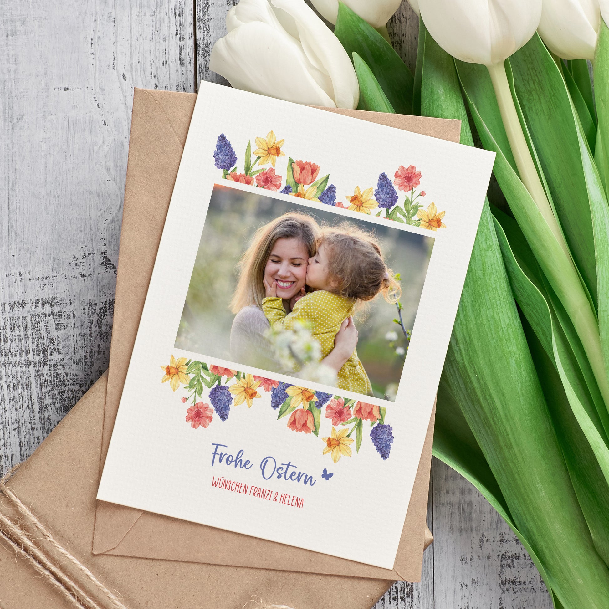 Osterkarte mit Foto und Blumenmotiv neben Umschlag und Tulpen dekorativ arrangiert