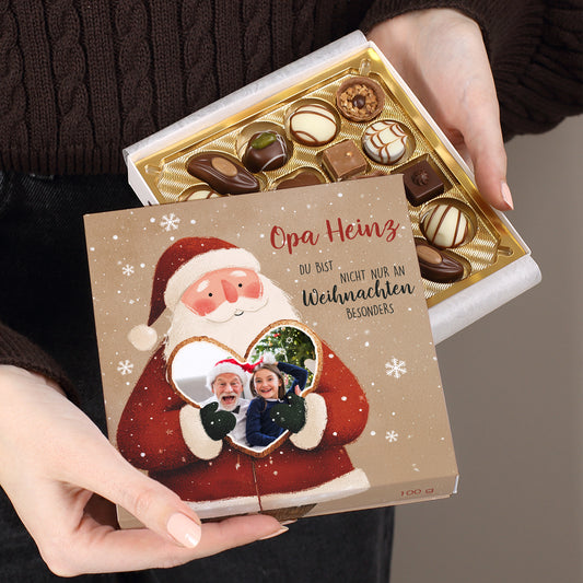 Weihnachtsmann-Motiv mit Foto eines Großvaters und eines Mädchens im Herz. Oben roter Schriftzug „Opa Heinz“, daneben schwarzer Wunschtext. Im Inneren goldene Pralinenauslage mit verschiedenen Formen und Dekoren.
