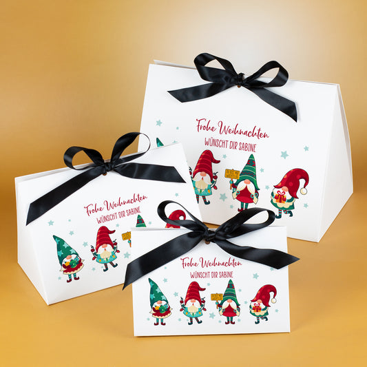Drei Geschenkverpackungen zu Weihnachten in den Größen klein, mittel und groß, jeweils aus mattweißem Karton mit schwarzem Satinband. Auf der Vorderseite ist ein farbiges Wichtelmotiv mit vier unterschiedlichen Wichteln zu sehen – einer mit Geschenken, einer mit Sternstab, einer mit Schild „Du bist mir wichtelig“ und einer mit Geschenkpaket. Jede Tasche trägt einen personalisierten roten Wunschtext. Die Taschen stehen auf goldenem Hintergrund und zeigen die Größenunterschiede deutlich.