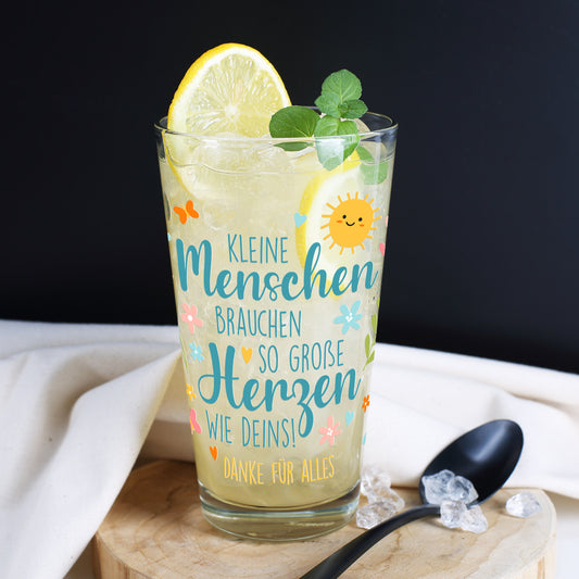 Nahaufnahme eines personalisierbaren Trinkglases mit transparenter Zitronenlimonade, dekoriert mit einer Zitronenscheibe und Minzblättern. Das Glas steht auf einem runden Holzbrett vor dunklem Hintergrund. Bedruckt mit dem Spruch „Kleine Menschen brauchen so große Herzen wie deins!“ sowie dem Zusatz „Danke für alles“. Das farbenfrohe Motiv zeigt eine lachende Sonne, Blumen, Herzen, Schmetterlinge und Blätter.