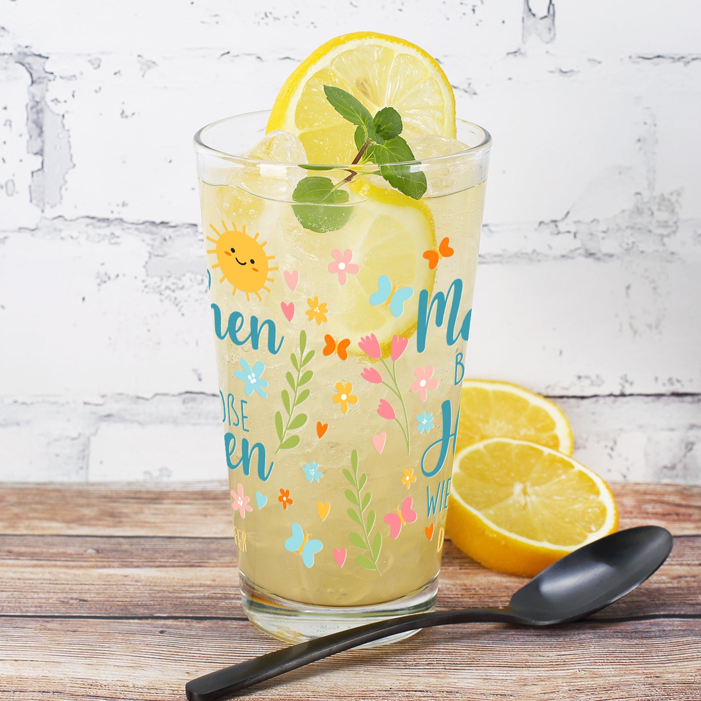 Trinkglas mit gelber Limonade, Zitronenscheibe und Minze, stehend auf einem Holztisch vor einer weiß gestrichenen Ziegelwand. Die Rückseite des Glases zeigt ein buntes Frühlingsmotiv mit Blüten, Schmetterlingen, Herzen und Zweigen. Die Vorderseite des Textes ist seitlich zu sehen.