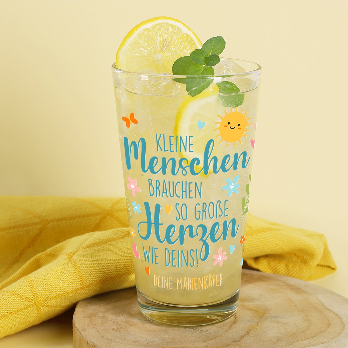 Trinkglas mit gelber Limonade, Zitrone und Minzblättern, arrangiert auf einem Holzbrett mit gelbem Handtuch im Hintergrund. Aufschrift in Türkis, Orange und Rosa: „Kleine Menschen brauchen so große Herzen wie deins!“ mit personalisiertem Wunschtext darunter: „Deine Marienkäfer“. Motiv mit Sonne, Herzchen, Schmetterlingen und Blüten.
