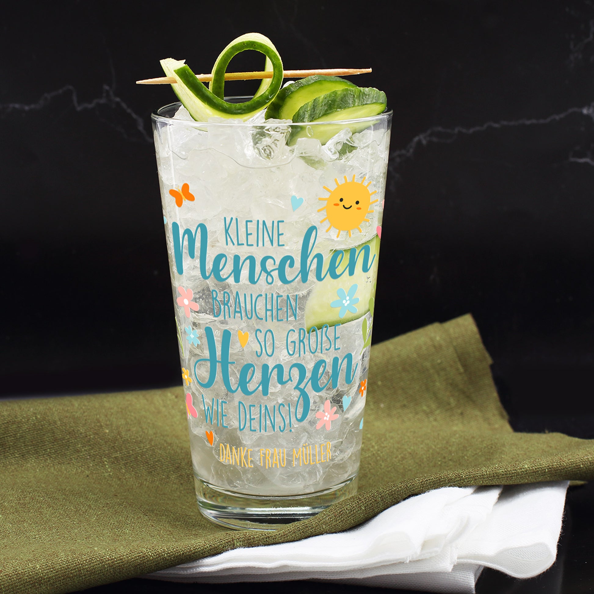 Trinkglas mit Gurkenlimonade und Eiswürfeln auf einem dunklen Untergrund. Garniert mit aufgerollter Gurkenspirale und dekorativem Holzspieß. Bedruckt mit dem Spruch „Kleine Menschen brauchen so große Herzen wie deins!“ und dem personalisierten Zusatz: „Danke Frau Müller“. Verspieltes Frühlingsmotiv mit lachender Sonne, Schmetterlingen, Blüten und bunten Elementen.