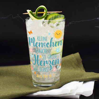 Trinkglas mit Gurkenlimonade und Eiswürfeln auf einem dunklen Untergrund. Garniert mit aufgerollter Gurkenspirale und dekorativem Holzspieß. Bedruckt mit dem Spruch „Kleine Menschen brauchen so große Herzen wie deins!“ und dem personalisierten Zusatz: „Danke Frau Müller“. Verspieltes Frühlingsmotiv mit lachender Sonne, Schmetterlingen, Blüten und bunten Elementen.