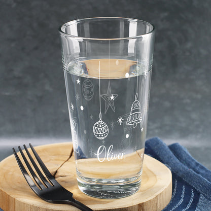Personalisierbares Weihnachtsglas mit weißen Kugel-, Stern- und Glockenmotiven, dekoriert mit Wasserfüllung und Namen, auf rustikalem Holzuntergrund.