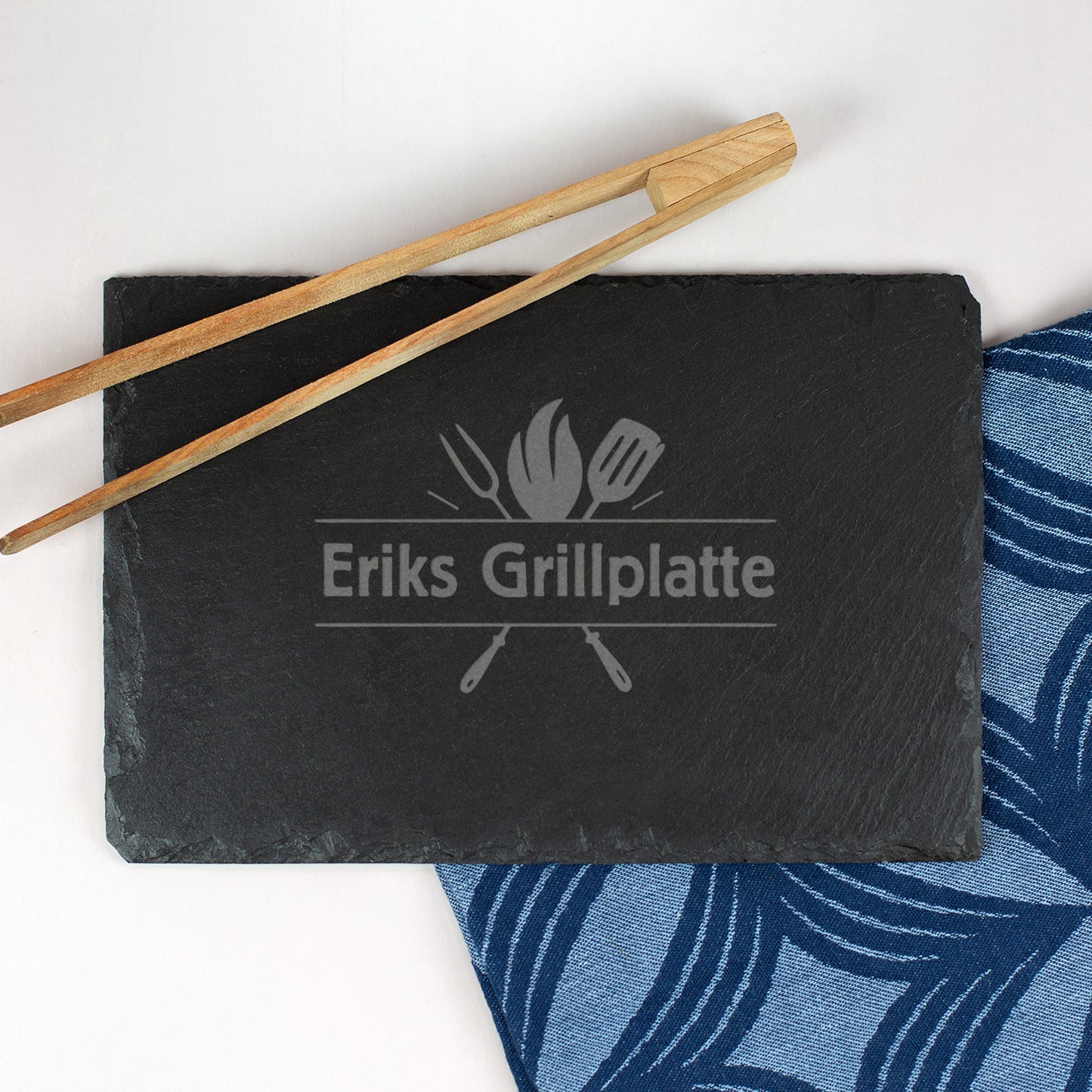 Schwarze Schieferplatte mit Gravur „Eriks Grillplatte“ und Grillutensilien, Holz-Grillzange als Dekoration