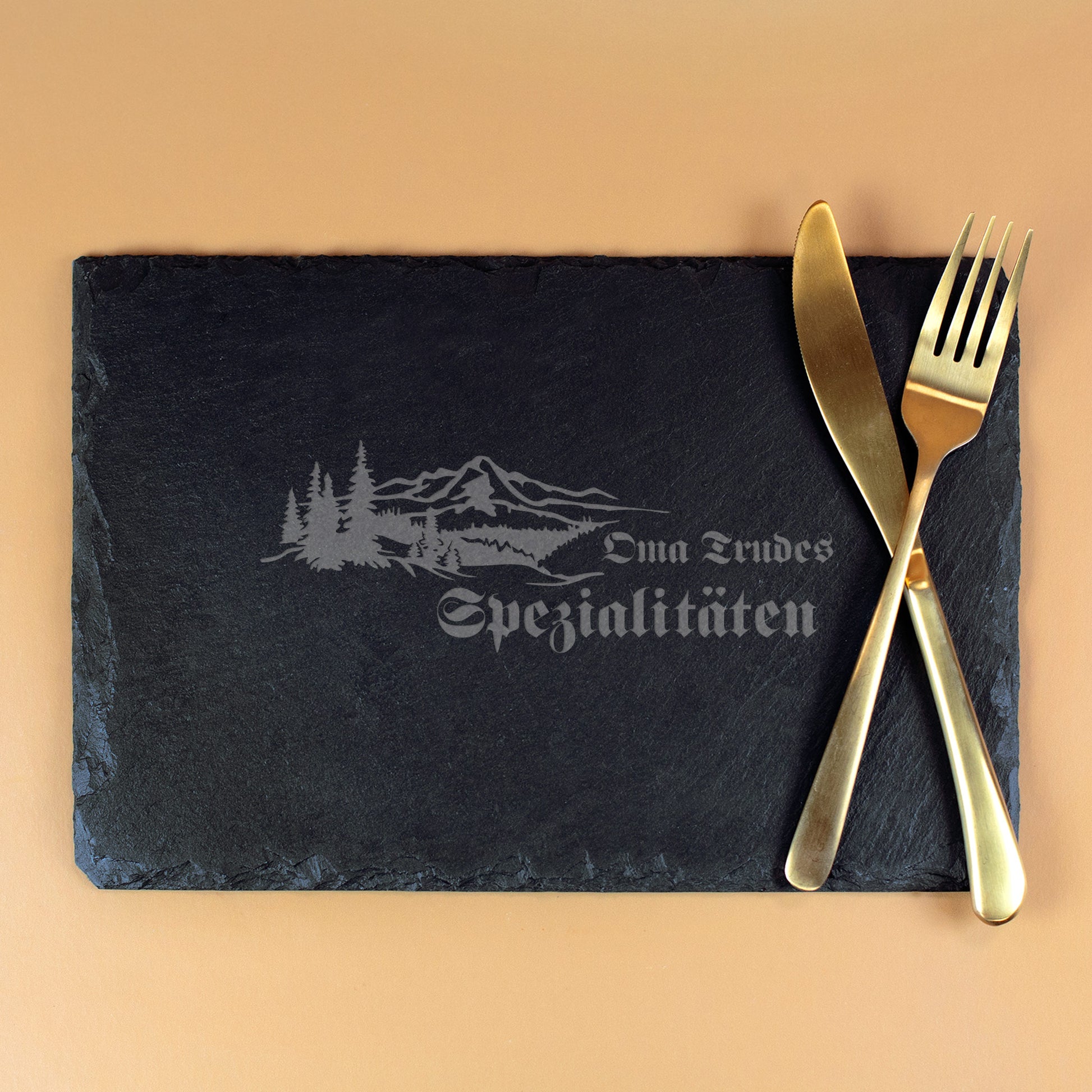 Schwarze Schieferplatte 30x20 cm mit Gravur „Oma Trudes Spezialitäten“ und Berg-Wald-Motiv, dekoriert mit goldenem Besteck.