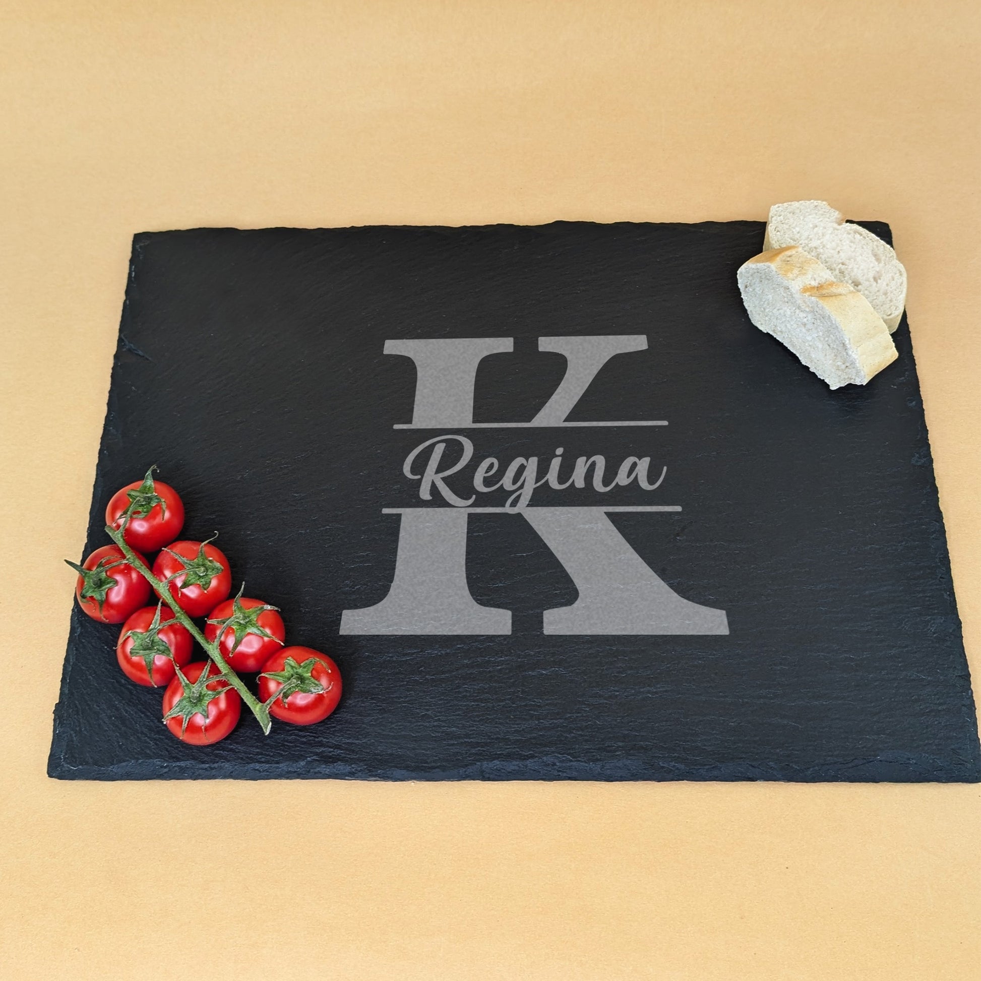 Schieferplatte mit Gravur „Regina“ und Initiale „K“, dekoriert mit Tomatenrispe und Brotscheiben auf beigem Hintergrund