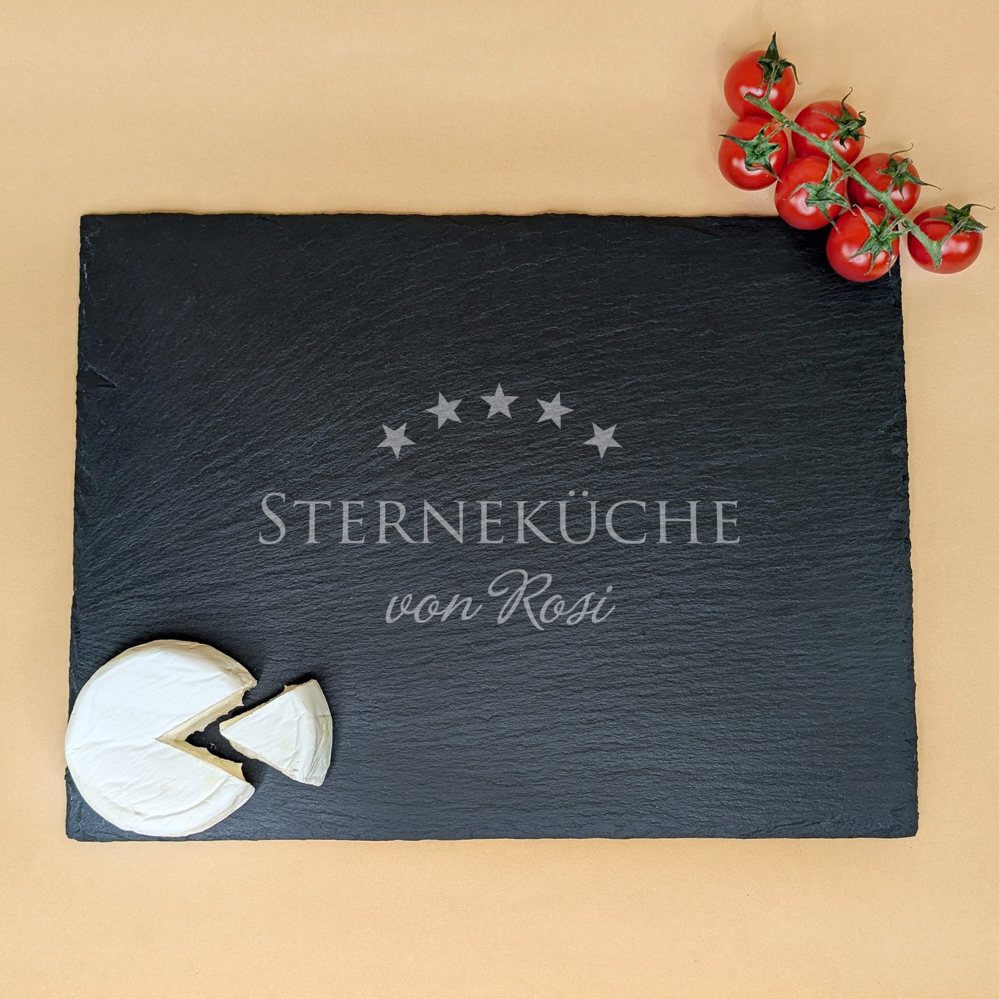 Schieferplatte mit Gravur „STERNEKÜCHE von Rosi“, mit Camembert und Tomatenzweig dekoriert – rustikaler Look auf beigem Hintergrund