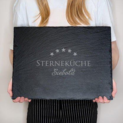 Schieferplatte mit Gravur „STERNEKÜCHE Siebold“, von einer Person in weißem Shirt und gestreifter Hose vor dem Körper gehalten – zur Größenansicht