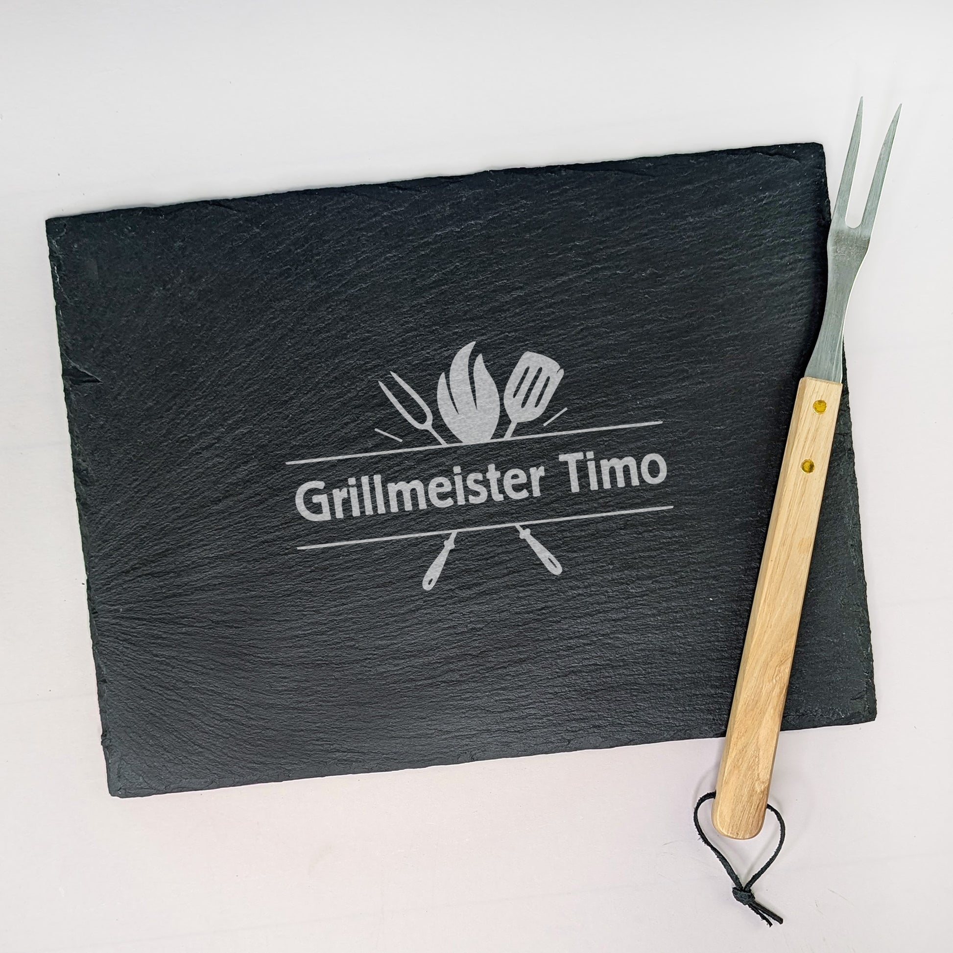 Schieferplatte mit Gravur „Grillmeister Timo“, daneben Grillgabel mit Holzgriff – fotografiert auf hellem Untergrund