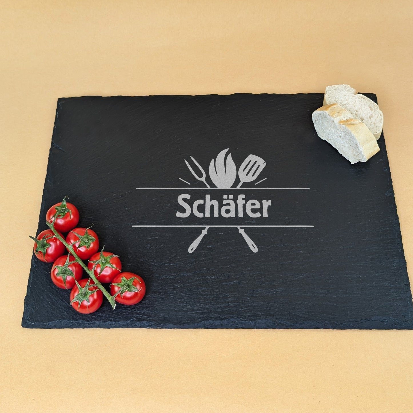 Schieferplatte mit Gravur „Schäfer“ und Grillmotiv, dekoriert mit Baguette und Tomatenrispe – angerichtet auf beigem Hintergrund