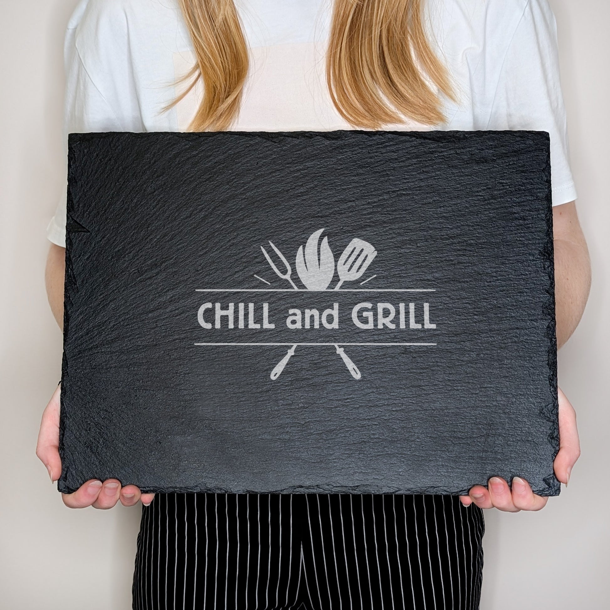 Schieferplatte mit Gravur „CHILL and GRILL“, in Händen gehalten zur Größenansicht, Person im weißen Shirt