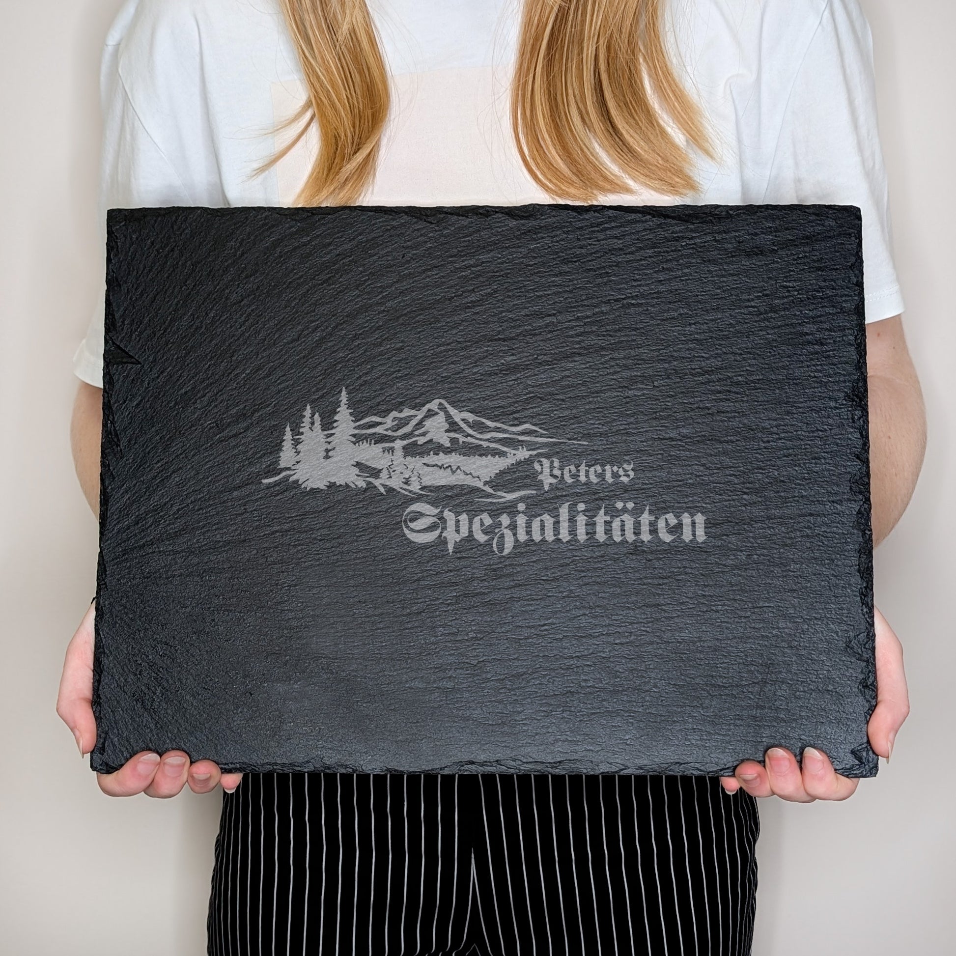 Rechteckige Schieferplatte mit Gravur „Peters Spezialitäten“, gehalten von einer Person in weißem Shirt