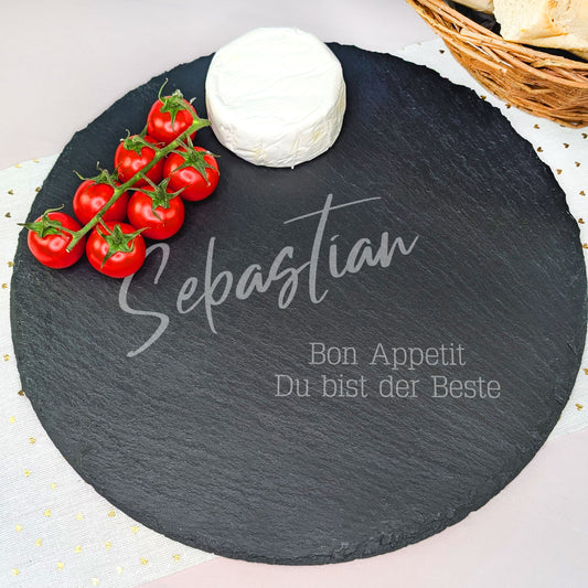 Runde Schieferplatte mit Gravur des Namens und einem zweizeiligen Spruch, dekoriert mit Camembert, Tomaten und Baguette-Korb.