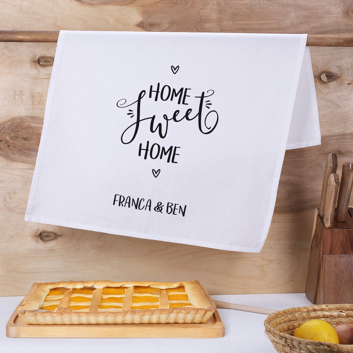 Bedrucktes Handtuch mit Motiv „Home sweet Home“ am Holzregal, darunter Kuchen auf Schneidebrett