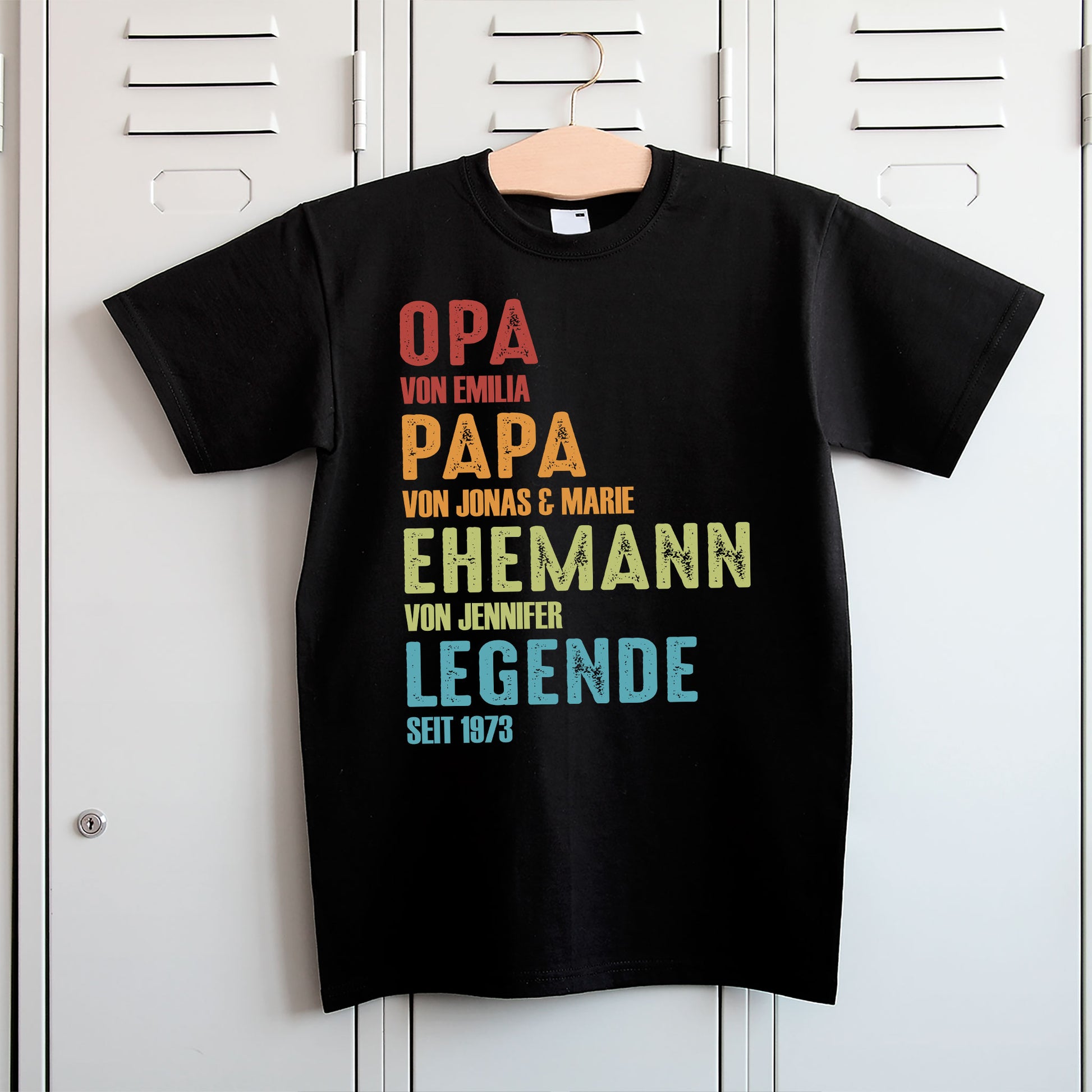 Schwarzes Herren-T-Shirt mit buntem Aufdruck „Opa seit … – Papa seit … – Ehemann seit … – Legende seit …“, hängend an Kleiderhaken fotografiert.