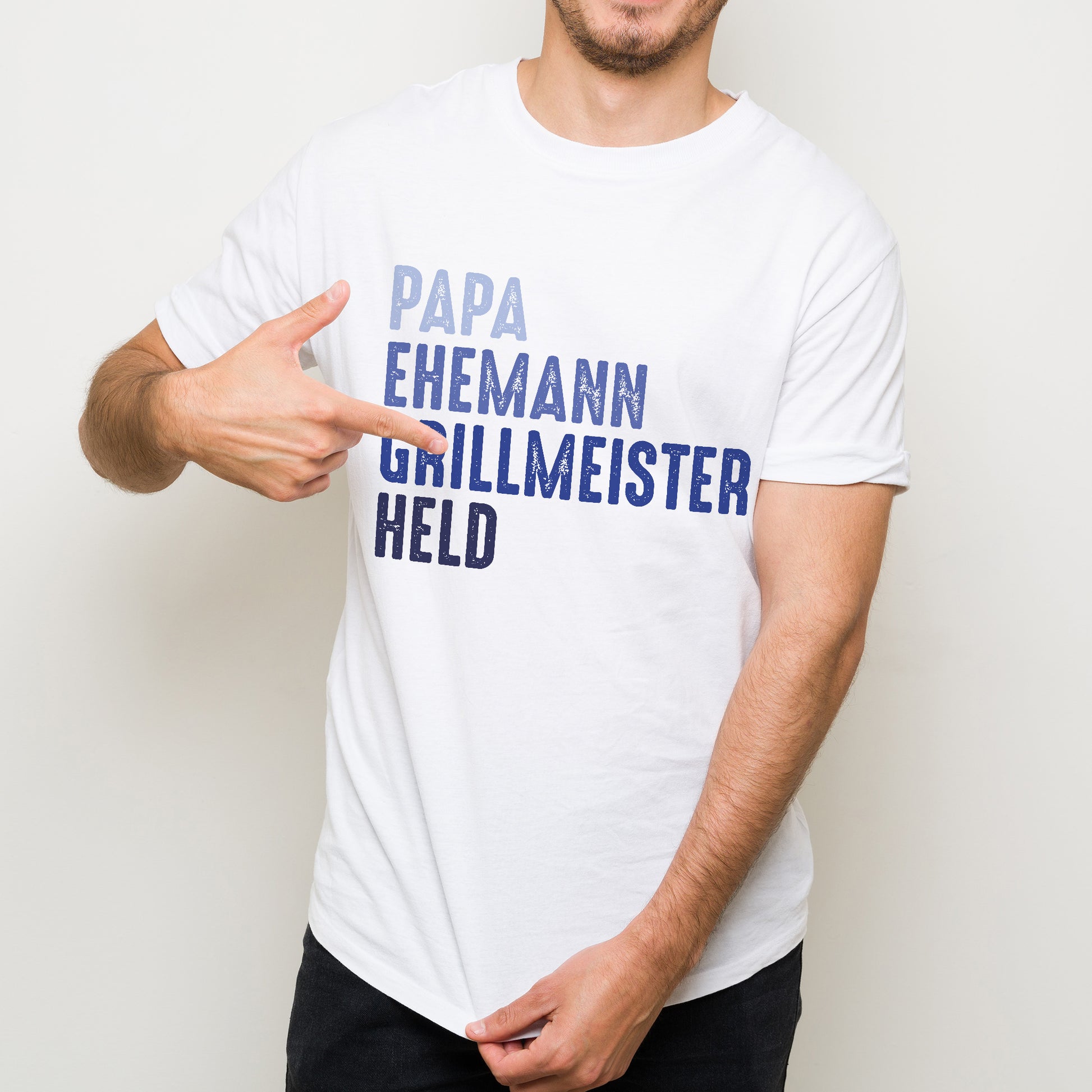 Mann zeigt auf sein weißes T-Shirt mit den Worten „Papa – Ehemann – Grillmeister – Held“