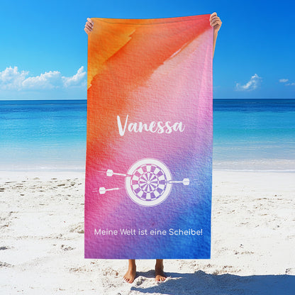 Person hält ein großes personalisiertes Badetuch mit Namen, Wunschtext und Hobby-Motiv am Strand in die Höhe – Druckmotiv gut sichtbar vor maritimer Kulisse.