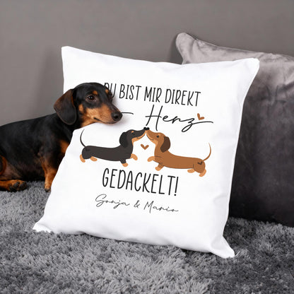 Personalisiertes Kissen mit zwei Dackeln und Herzchen, Spruch „Ins Herz gedackelt!“, Hund liegt daneben auf Sofa