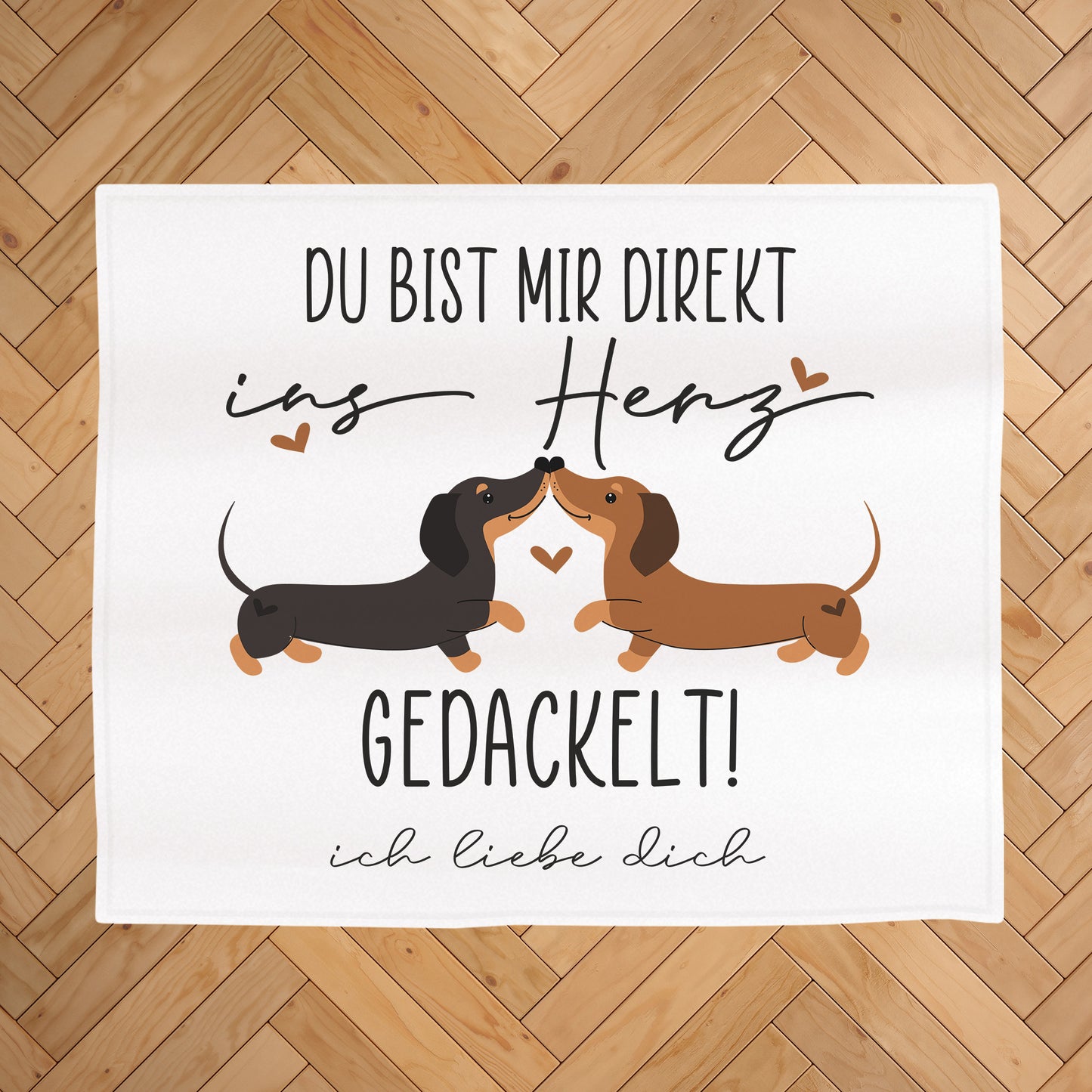 Kuscheldecke mit Dackel-Motiv auf hellem Holz-Fischgrätboden, Aufdruck „Du bist mir direkt ins Herz gedackelt!“ und Wunschtext „ich liebe dich“ unter dem Motiv