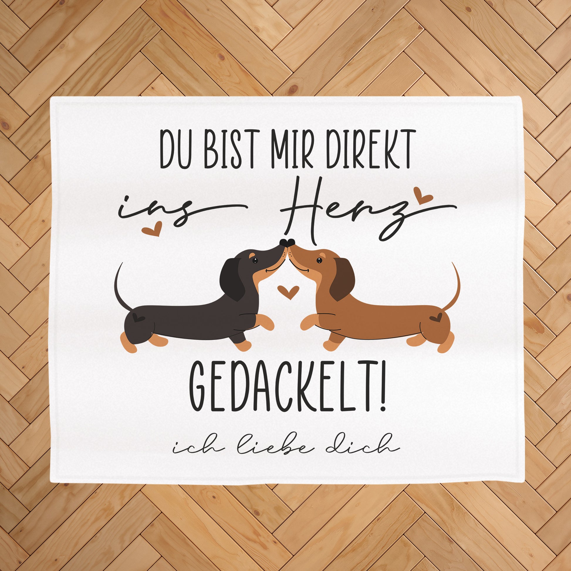 Kuscheldecke mit Dackel-Motiv auf hellem Holz-Fischgrätboden, Aufdruck „Du bist mir direkt ins Herz gedackelt!“ und Wunschtext „ich liebe dich“ unter dem Motiv