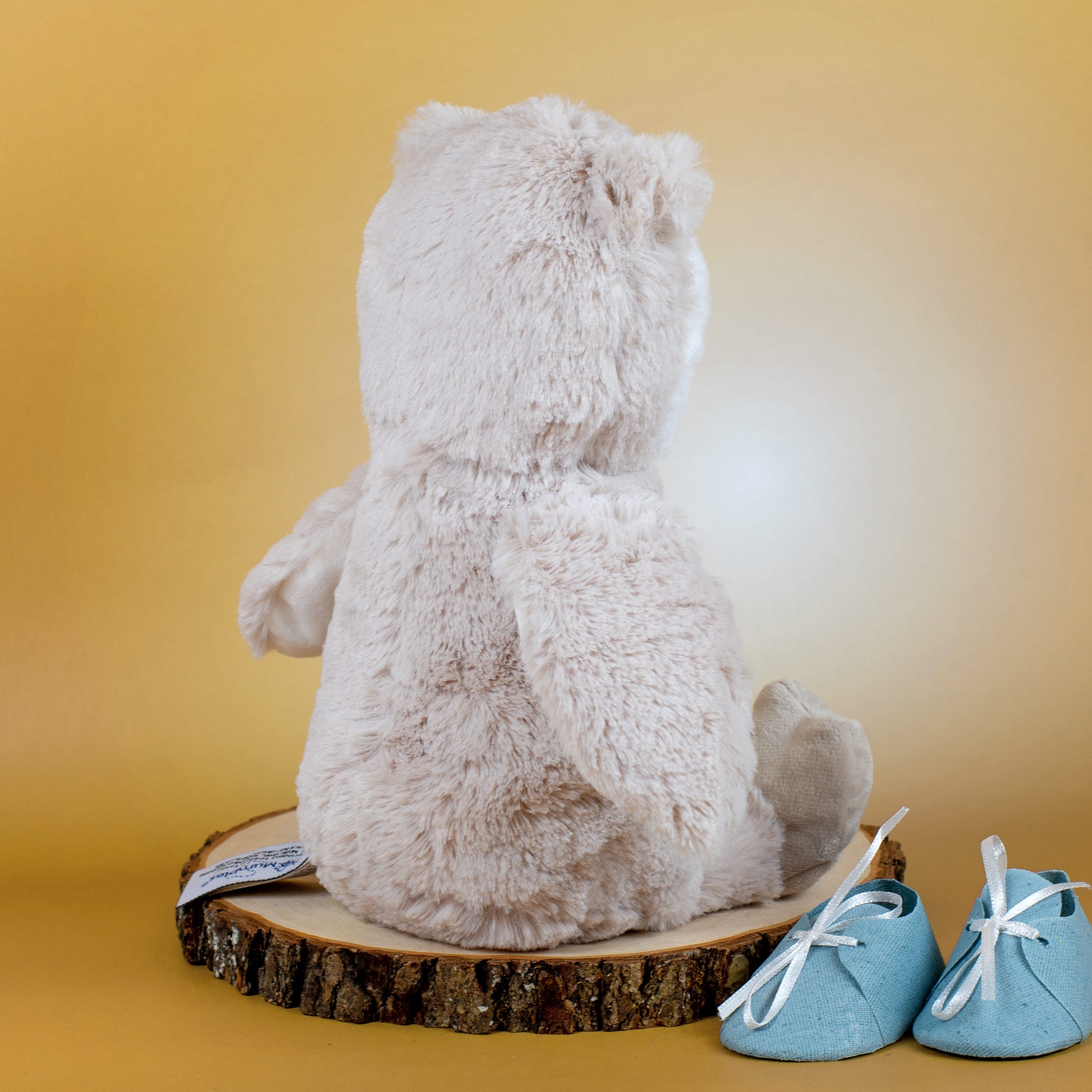 Rückansicht einer flauschigen, beigefarbenen Kuscheltier-Eule auf einer Holzscheibe, daneben hellblaue Babyschuhe mit weißen Bändern.