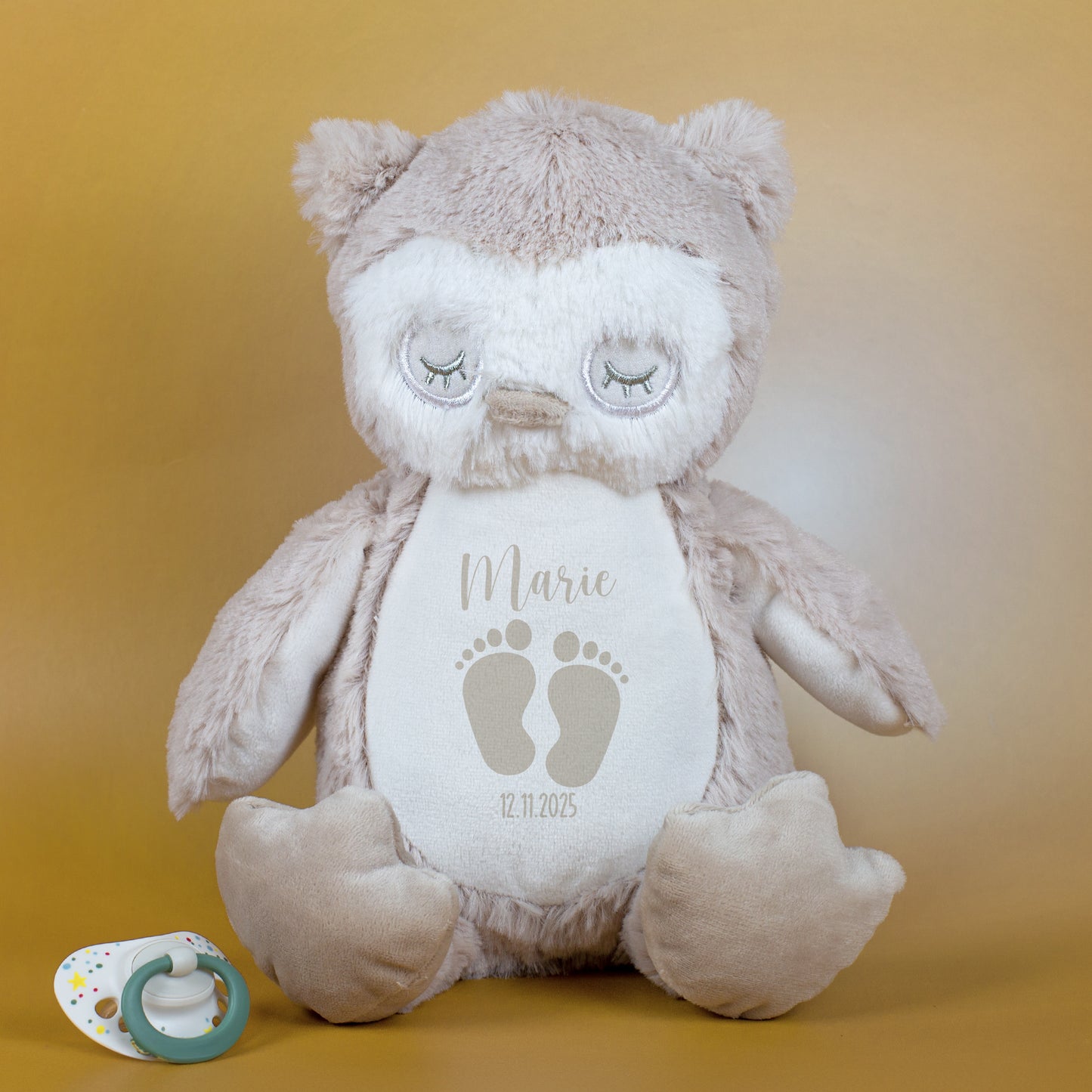 Eine beige Eule aus Plüsch mit besticktem Gesicht und geschlossenen Augen sitzt auf einer goldfarbenen Fläche. Auf dem Bauch steht in grauer Schrift der Name „Marie“ und das Geburtsdatum „12.11.2025“ unter einem Paar Babyfüßchen.
