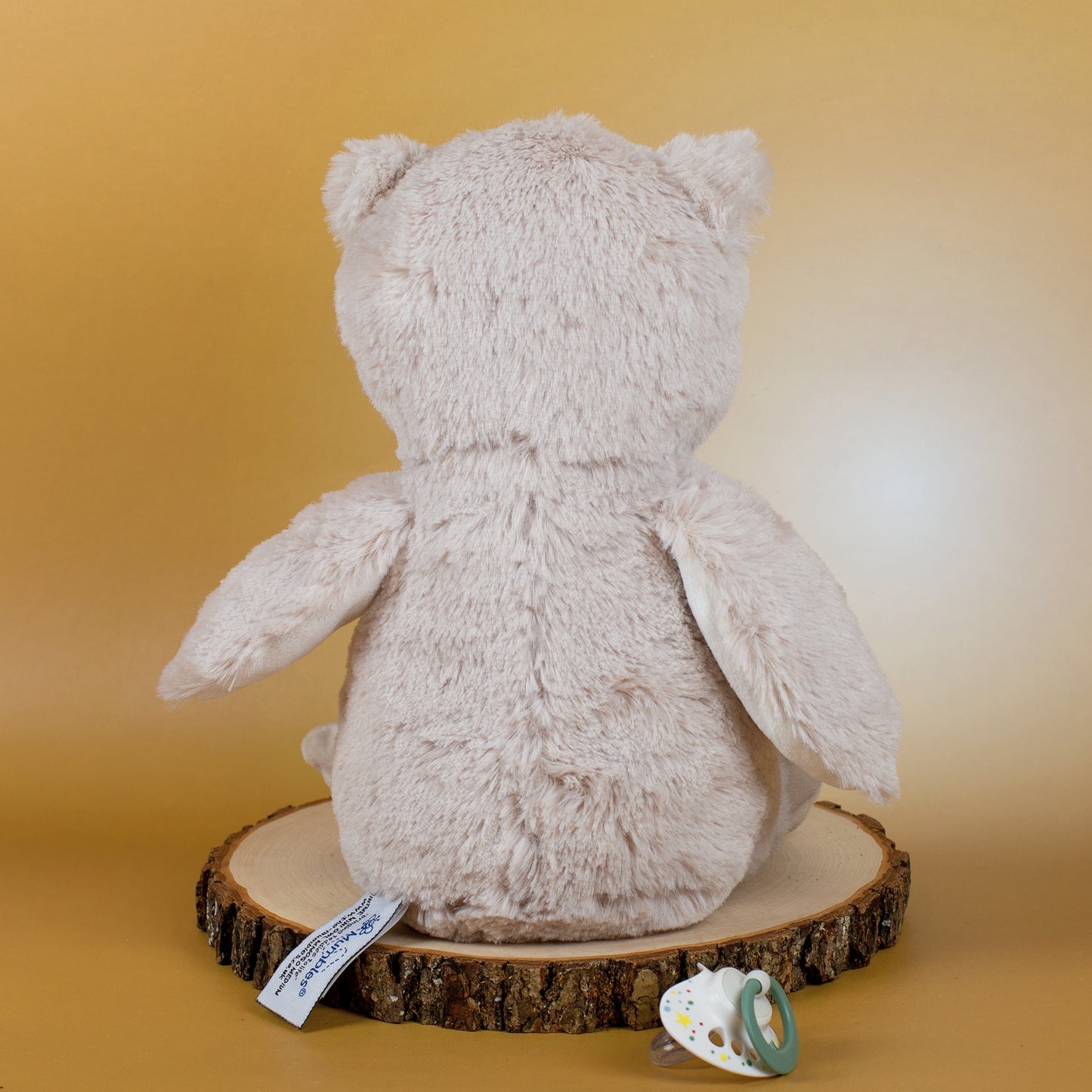 Die Eule ist von hinten zu sehen, sie sitzt auf einer Holzscheibe. Daneben liegt ein grüner Schnuller, der das Foto als Geschenkidee für Babys unterstreicht.