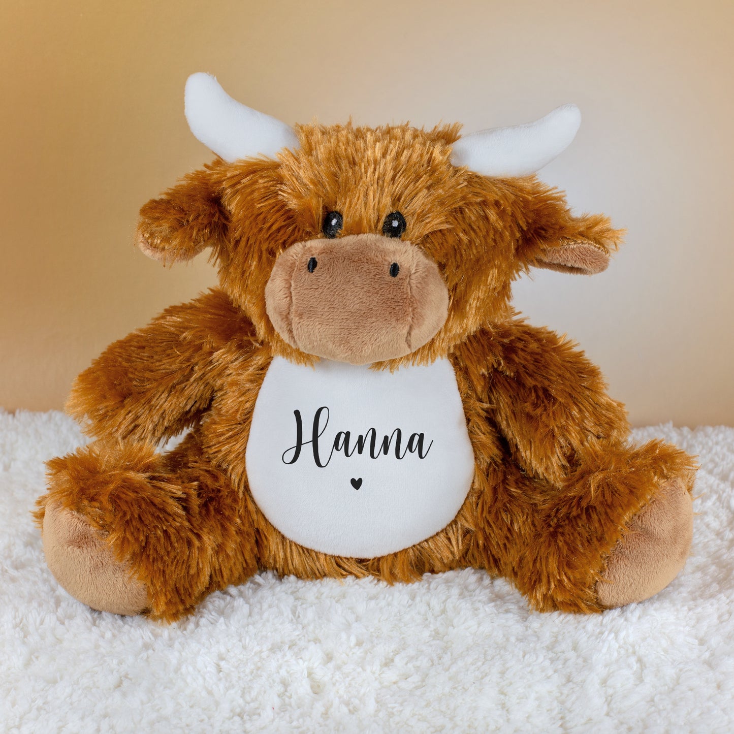 Personalisierte Kuscheltier-Kuh mit Name Hanna über kleinem Herzmotiv auf weißem Bauch, sitzend auf weicher Decke.