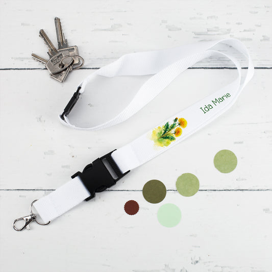 Weißes Schlüsselband mit Löwenzahn-Motiv und dem Namen "Ida Marie", neben einem Schlüsselbund auf weißem Untergrund