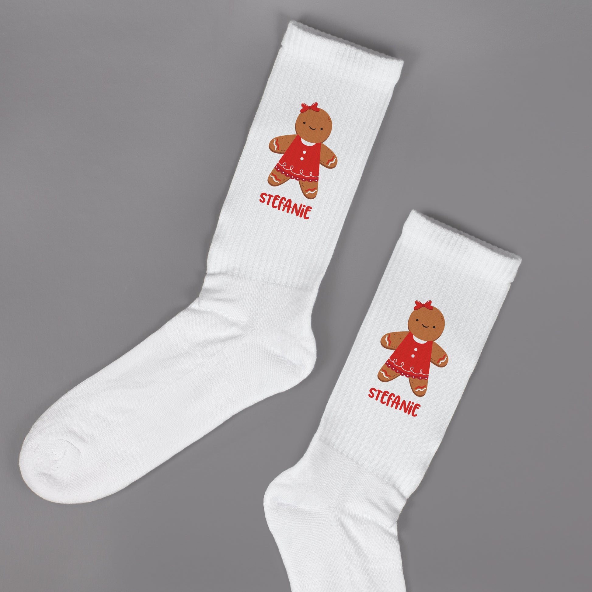 Paar weißer Socken mit Lebkuchenfrau-Motiv und personalisiertem Namen, flach auf grauer Oberfläche.