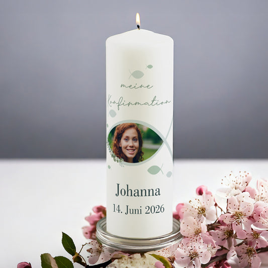 Personalisierte Kerze zur Konfirmation mit Foto. Die weiße Kerze zeigt den Schriftzug „meine Konfirmation“, ein christliches Fischsymbol mit integriertem Foto eines Mädchens und darunter Name „Johanna“ mit Datum „14. Juni 2026“. Die Kerze steht angezündet auf einem Glasteller, umgeben von rosa Blüten.