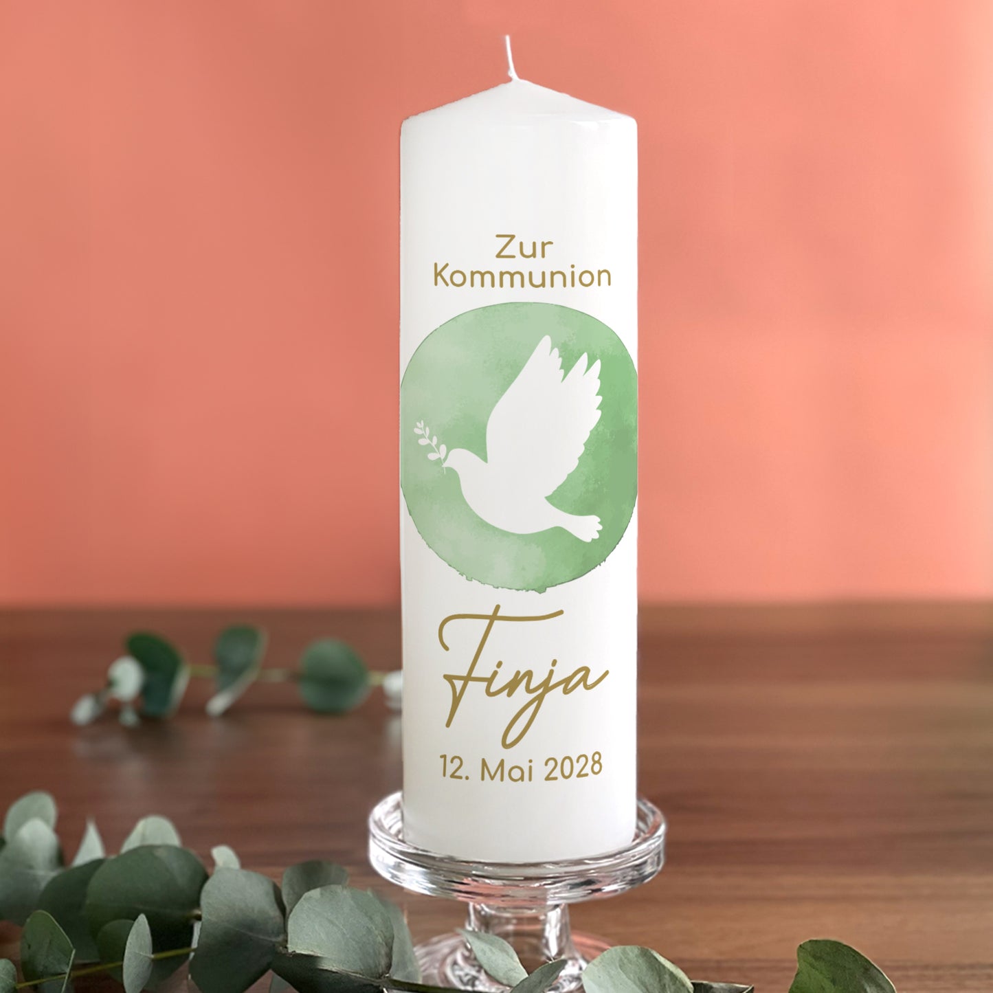 Personalisierte Kerze zur Kommunion mit grünem Aquarellmotiv einer Taube mit Zweig. Der Name "Finja" und das Datum "12. Mai 2028" sind in goldener Schrift ergänzt. Die festliche Kerze steht auf einem Kerzenhalter, umgeben von Eukalyptusblättern, vor einem warmen orangefarbenen Hintergrund.