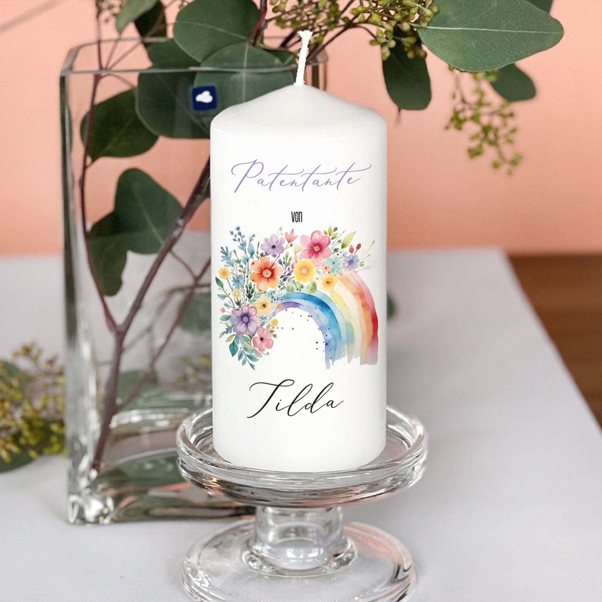 Patenkerze mit Regenbogenmotiv, Text „Patentante von Tilda“, platziert auf Glasständer mit Eukalyptus im Hintergrund