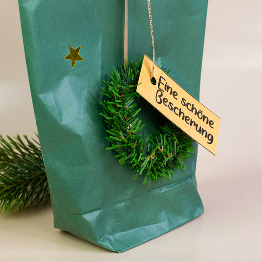 Nahaufnahme Dekokranz mit Anhänger „Eine schöne Bescherung“ an der grünen Geschenktüte
